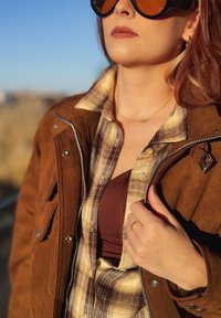 Giacca di suede marrone indossata sopra una camicia a quadri marrone e beige, abbinata a una maglia bordeaux. Grandi occhiali da sole neri, anello delicato sulla mano.