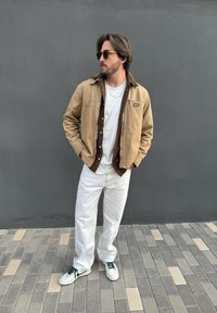 Beżowo-brązowa warstwowa kurtka, biały T-shirt i białe spodnie. Sneakersy z zielonymi akcentami. Szara ściana i brukowana ziemia w tle.