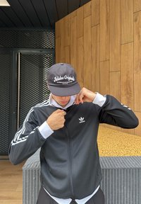 Fekete Adidas cipzáras dzseki fehér csíkokkal az ujjakon, fehér póló fölé húzva. Fekete sapka "Adidas Originals" logóval. Modern háttér.