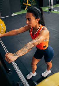 Svara stienis ar dzeltenām svaru plāksnēm; indivīds ar sarkanu sporta kreklu un tumši ziliem šortiem tur stieni, uz viņa rokas ir redzams detalizēts ziedu tetovējums.