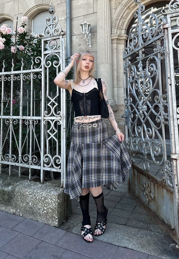 Jeune personne avec des tatouages porte un top en dentelle noire, une jupe à carreaux et des sandales avec des chaussettes transparentes montant aux genoux, debout près d'une porte métallique ornée et d'un mur en pierre.