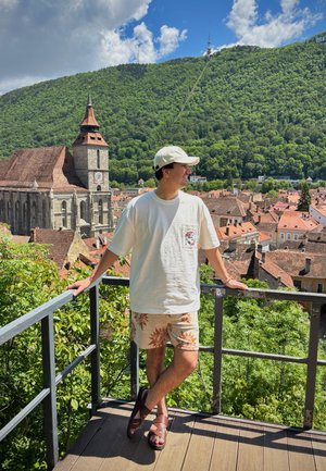 Conjunto informal que incluye una camiseta blanca, pantalones cortos con un patrón en beige con diseños florales marrones, y sandalias marrones, todo en un fondo de montañas verdes.