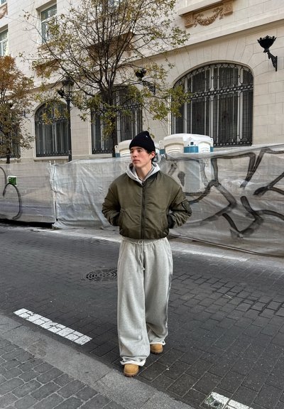 Chaqueta bomber verde oliva, sudadera gris claro, pantalones de chándal grises holgados y botas marrón claro. Fondo urbano con árboles y barreras de construcción.