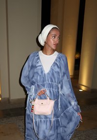 Μπλε tie-dye maxi φόρεμα με μακριά μανίκια και ρυθμιζόμενη μέση. Κρατάει μια μικρή ροζ τσάντα χειρός με διακοσμητικό γούρι. Λευκό μαντίλι.