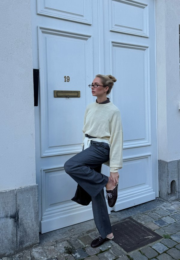 Mujer con gafas que lleva un suéter crema y pantalones grises, de pie sobre una pierna, ajustándose el zapato oscuro delante de una puerta blanca con el número 19.