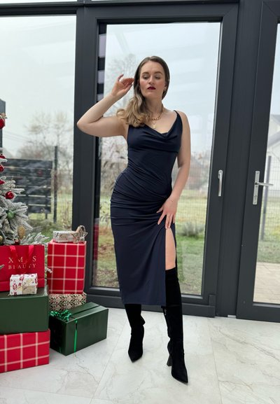 Vestido azul marino con cuello en cowl, fruncidos y una abertura hasta el muslo, combinado con botas negras hasta la rodilla, en una habitación decorada con regalos.