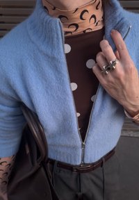 Pull à fermeture éclair bleu clair avec tricot texturé, superposé à un haut marron à pois. Bague étoile en argent et sac à main marron visibles.