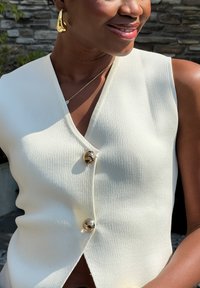 Ανοιχτό κρεμ αμάνικο τοπ με βαθύ V ντεκολτέ, που διαθέτει υφή ριμπ και δύο μεγάλους χρυσούς κουμπιά μπροστά. Απλός, μινιμαλιστικός σχεδιασμός.