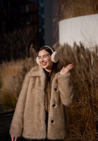 Femme souriante portant un manteau en fausse fourrure beige et des cache-oreilles duveteux, qui fait signe en se tenant à l'extérieur avec de hautes herbes sèches en arrière-plan.