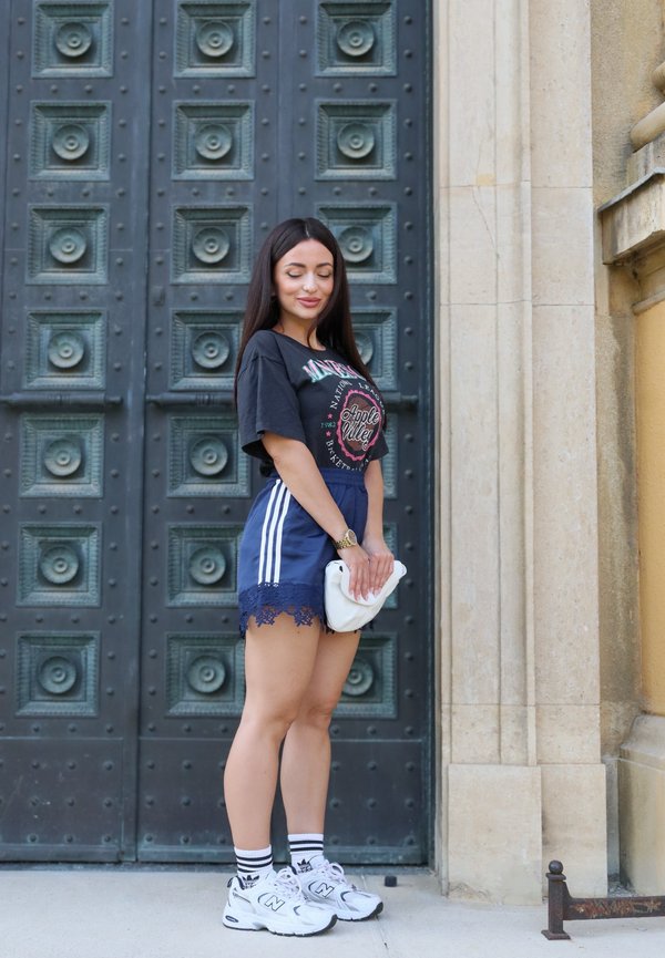 Kvinde med langt mørkt hår står foran en stor udsmykket metal dør iført marineblå shorts, sort grafisk t-shirt, hvide sneakers og holder en hvid clutch.