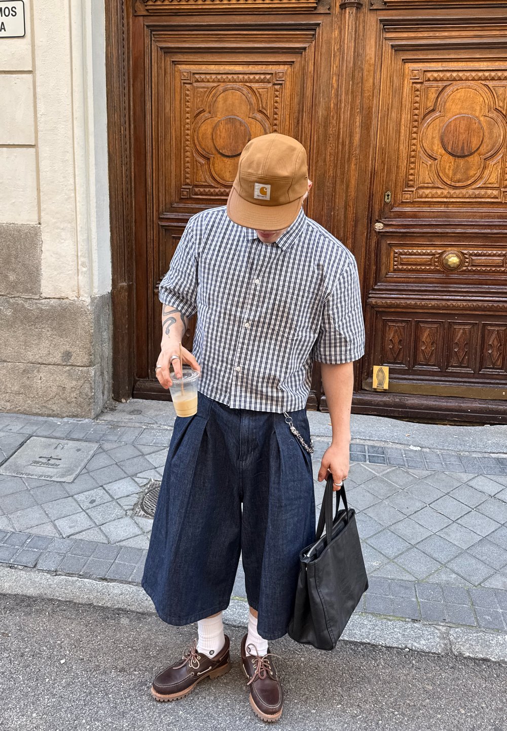 Kratkoročna karirana srajca, temno modri culottes, rjava kapa, rjave čevlje, z verižnim detajlom in v rokah pijačo, črna torba.