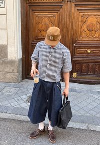 Kratkoročna karirana srajca, temno modri culottes, rjava kapa, rjave čevlje, z verižnim detajlom in v rokah pijačo, črna torba.