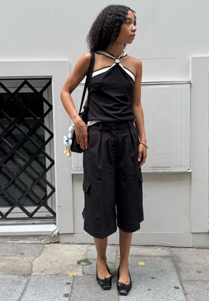 Mujer con pelo rizado que lleva un top halter negro y pantalones cortos negros largos, de pie en la acera de la ciudad sosteniendo un bolso negro de hombro.