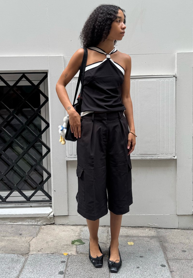 Donna con capelli ricci che indossa un top a halter nero e pantaloni lunghi neri, si trova sul marciapiede della città tenendo una borsa a spalla nera.
