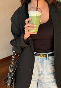 Blazer oversized preto, top preto, jeans claros de cintura alta, cinto com fivela dourada, segurando uma bebida gelada verde com um canudo.