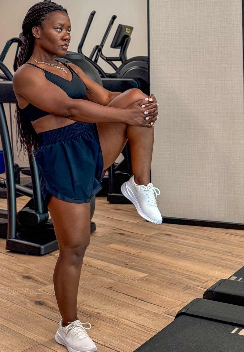 Femme en tenue de sport étirant une jambe tout en se tenant debout sur un sol en bois avec du matériel d'exercice en arrière-plan.
