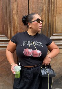 T-shirt preta com gráfico de basquetebol, saia preta, segurando um copo transparente com bebida verde, accessorized com óculos de sol e uma pequena bolsa preta.