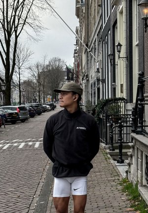 Sudadera de forro polar negra con cuello alto y logo, pantalones cortos ajustados blancos con el logo negro de Nike y gorra verde oliva, de pie en una calle adoquinada.