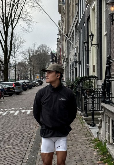 Sudadera de forro polar negra con cuello alto y logo, pantalones cortos ajustados blancos con el logo negro de Nike y gorra verde oliva, de pie en una calle adoquinada.