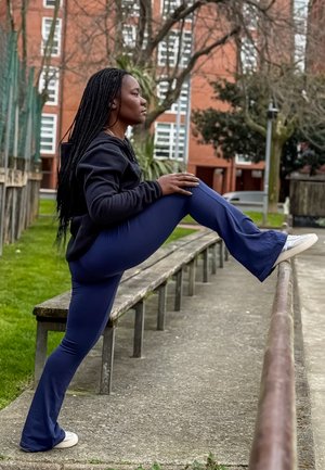 Mujer estirando la pierna sobre una barandilla metálica a lo largo de un sendero en un parque urbano cerca de bancos y edificios de ladrillo.