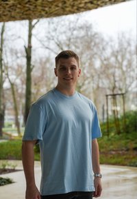Ljusblå t-shirt med kort ärm och rund halsringning, med broderad logga på bröstet, i kombination med mörka jeans och en silverfärgad klocka.