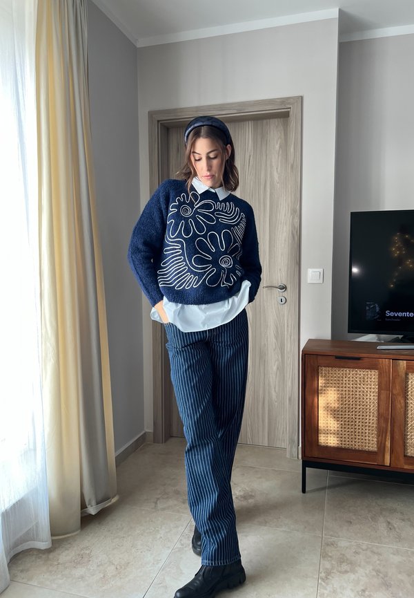 Marineblauer Strickpullover mit weißem Blumendesign, über einem weißen Hemd getragen. Gestreifte marineblaue Hose und schwarze Knöchelstiefel runden das Outfit ab.