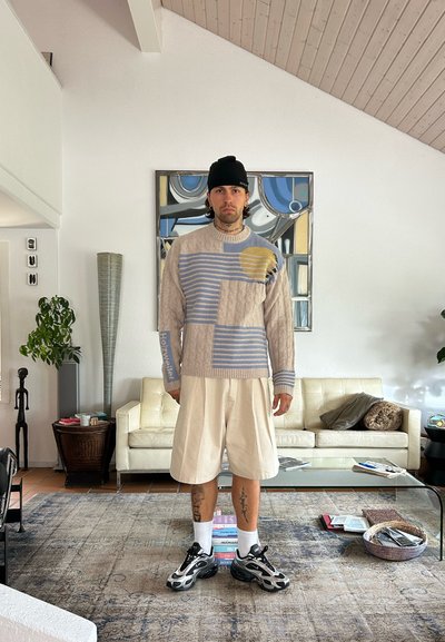 Suéter de punto con rayas beige y azules, acento amarillo. Combinado con pantalones cortos crema, zapatillas negras y calcetines blancos. Entorno interior con muebles.