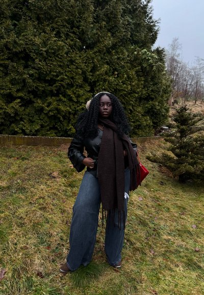 Mujer con cabello rizado, vestida con chaqueta de cuero negra, jeans anchos, una bufanda larga oscura y orejeras, de pie sobre el césped cerca de altos árboles perennes.