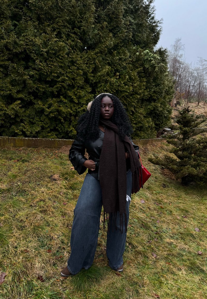 Femme aux cheveux bouclés portant une veste en cuir noire, un jean large, une longue écharpe sombre et des cache-oreilles, debout sur l'herbe près de hauts conifères.