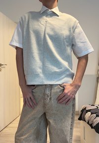 Top gri fără mâneci purtat peste o cămașă albă cu guler; asortat cu pantaloni bej deschis, cu textură subtilă și detalii de cusături.