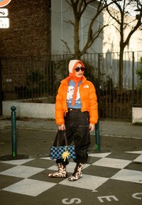Personne portant une doudoune orange vif, un t-shirt graphique bleu, un pantalon noir, des bottes imprimées vache et un chapeau blanc et orange, debout sur un passage piéton en tenant un sac.