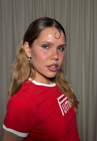 Ung kvinna med ljusblå ögon, vågigt blont hår, en lock på pannan, iklädd diamanthänge och en röd Puma-t-shirt, vänd något åt sidan.