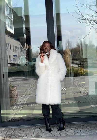 Abrigo de piel sintética blanca con cuello alto y mangas anchas, combinado con pantalones negros ajustados y botines de tacón alto negros.