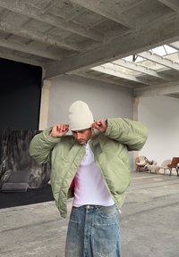 Bomber bunda olivově zelená se zipem, prošívaná textura, elastické manžety, nošena přes bílou henley košili, doplněná béžovou čepicí. Džínové kalhoty.