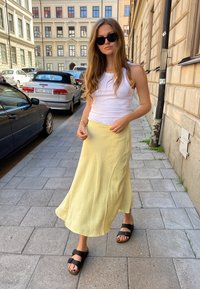 Sárga pöttyös szoknya, fehér topp, fekete napszemüveg és barna szandál. Hétköznapi utcai jelenet, macskaköves járdával.