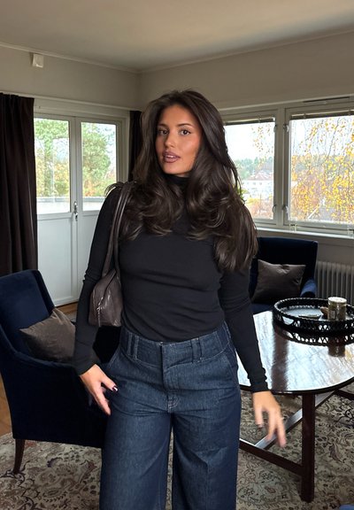 Mujer con cabello largo y oscuro, vestida con un jersey negro de cuello alto y vaqueros azules de talle alto, en una sala de estar con sillones y una mesa de madera.