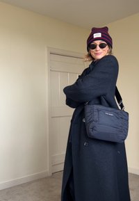 Personne portant un manteau noir, un bonnet rayé, des lunettes de soleil et un sac en bandoulière, debout à l'intérieur avec les bras croisés, souriant légèrement.