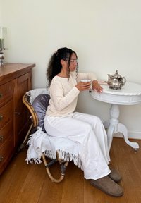 Suéter de malha creme, calças brancas de perna larga e calçado castanho. Sentada numa cadeira de madeira com encosto trançado, segurando uma chávena perto de uma mesa branca.