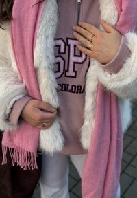Sciarpa rosa soffice indossata sopra una felpa rosa chiaro con la scritta "SP", circondata da un cappotto di pelliccia ecologica bianca con polsini e trama visibili.