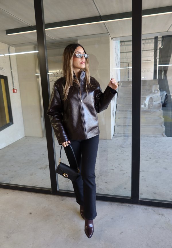 Frau trägt dunkelbraune Lederjacke, schwarze Hose, spitze Stiefel, Sonnenbrille, hält kleine schwarze Handtasche, steht vor einer Glastür.