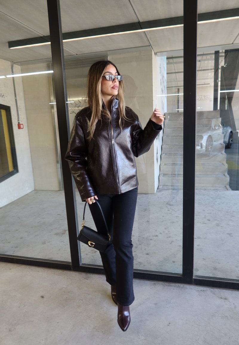 Frau trägt dunkelbraune Lederjacke, schwarze Hose, spitze Stiefel, Sonnenbrille, hält eine kleine schwarze Handtasche, steht vor einer Glastür.