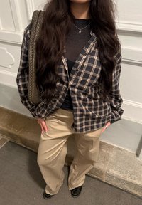 Femme aux longs cheveux foncés portant un haut en maille noire, une chemise à carreaux, un pantalon beige à jambes larges, des chaussures noires, portant un sac tissé à l'épaule.