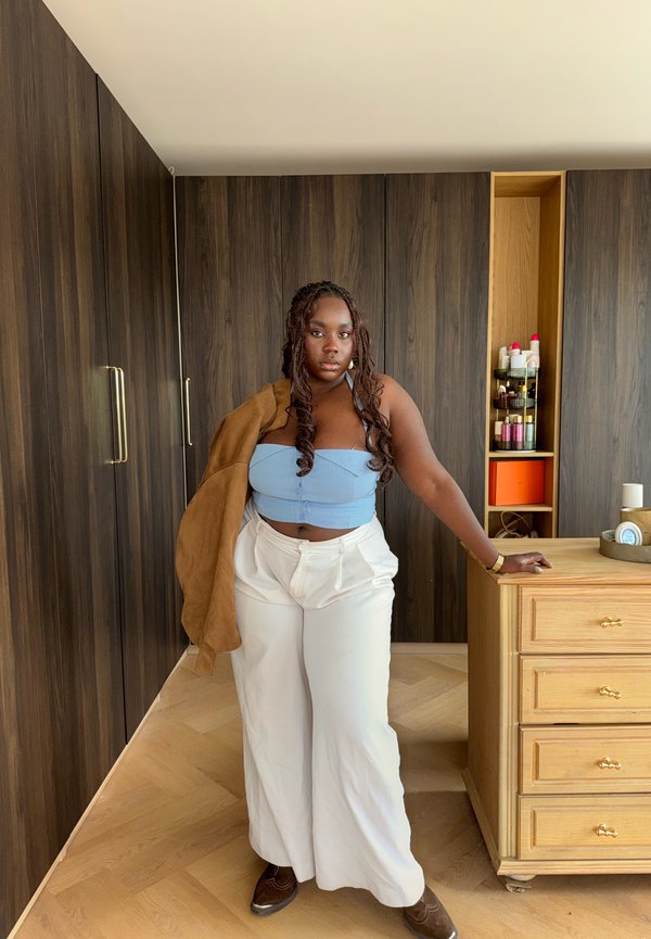 Vrouw met lang gevlochten haar die een blauw crop top, witte wijde broek en een bruine jas over één schouder gedrapeerd draagt in een kamer met houten panelen.