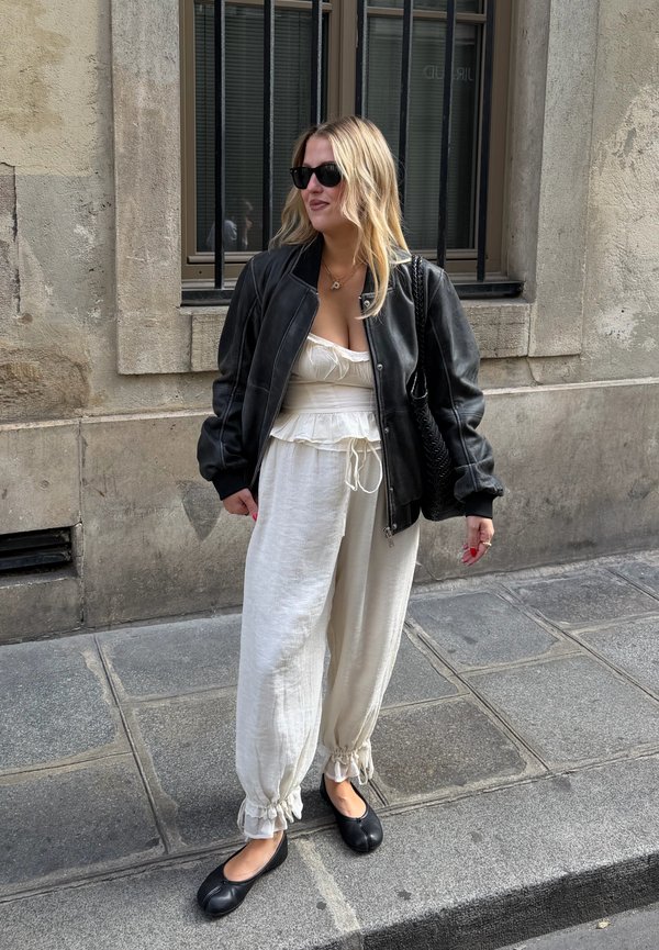 Une femme portant des lunettes de soleil, une veste en cuir noire, une tenue blanche et des chaussures plates se tient sur un trottoir en ville, à côté d’un bâtiment en pierre.