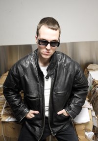 Chaqueta bomber de cuero negra con bolsillos laterales, llevada sobre una camisa blanca. La persona lleva gafas de sol oscuras y tiene el pelo corto.