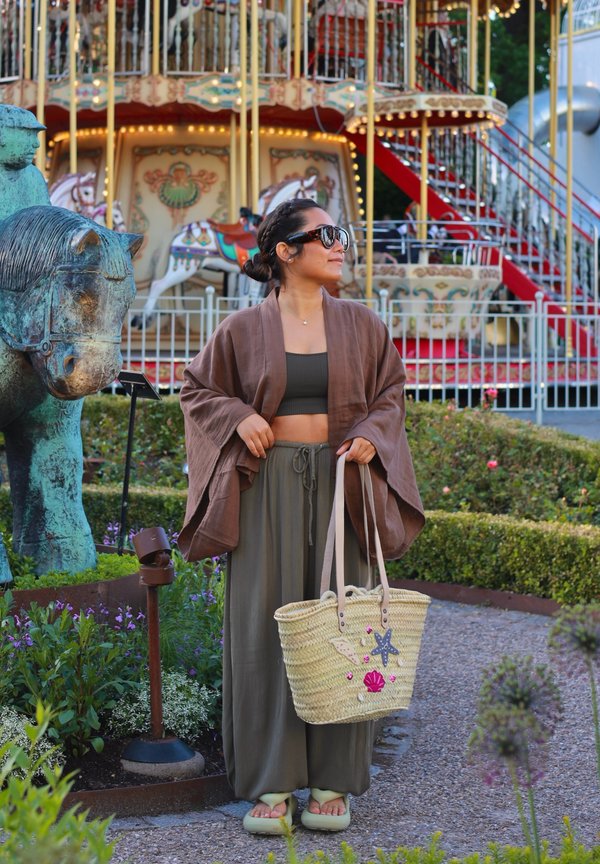 Frau mit Sonnenbrille und lockerer Kleidung steht nahe einem Karussell und einer grünen Pferdestatue und hält eine verzierte Strohtasche.