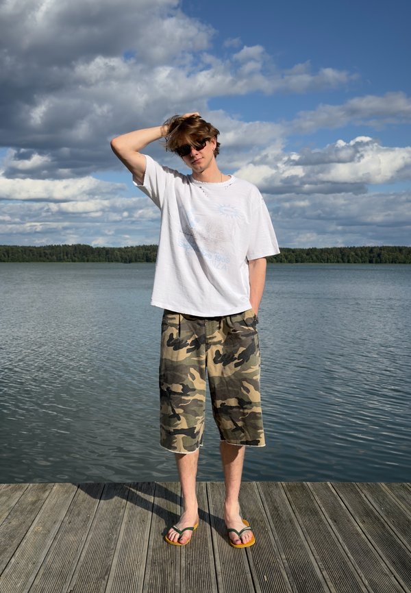 Un joven con gafas de sol, camiseta blanca, pantalones cortos de camuflaje y chanclas está de pie en un muelle de madera junto al lago bajo un cielo parcialmente nublado.