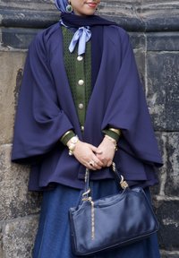 Marineblauwe cape-achtige outerwear, groene gestructureerde trui met knopen, denim rok en een marineblauwe handtas met een gouden kettingband. Elegante oorbellen en ringen.