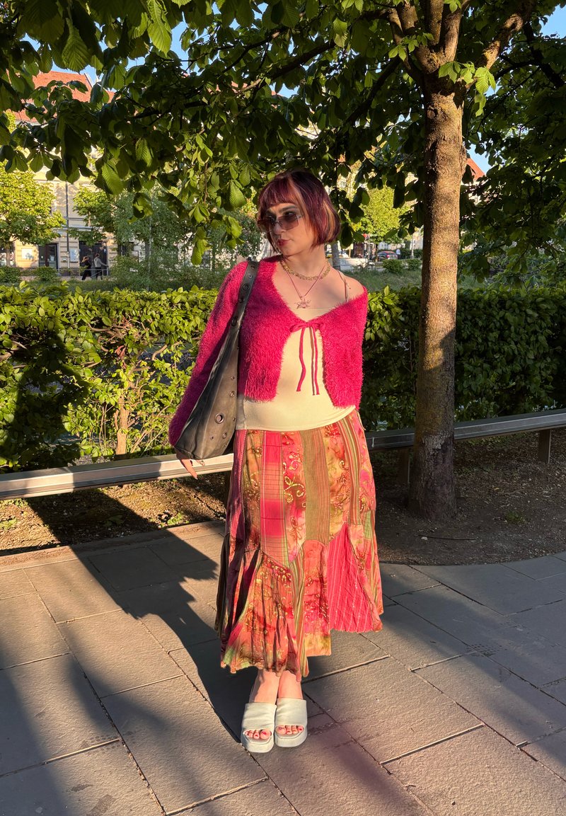 Jeune personne aux cheveux courts portant des lunettes de soleil, un cardigan rose, une longue jupe à motifs, des sandales blanches et portant un sac noir sous un arbre.