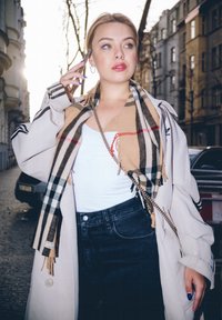 Béžový dlouhý trenčkot s černými pruhy, šátek s Burberry vzorem, bílý top, tmavé džíny a kabelka přes rameno na řetízku, držící telefon.